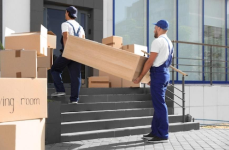 Two movers carrying a large box up steps outside a building, with stacked moving boxes nearby