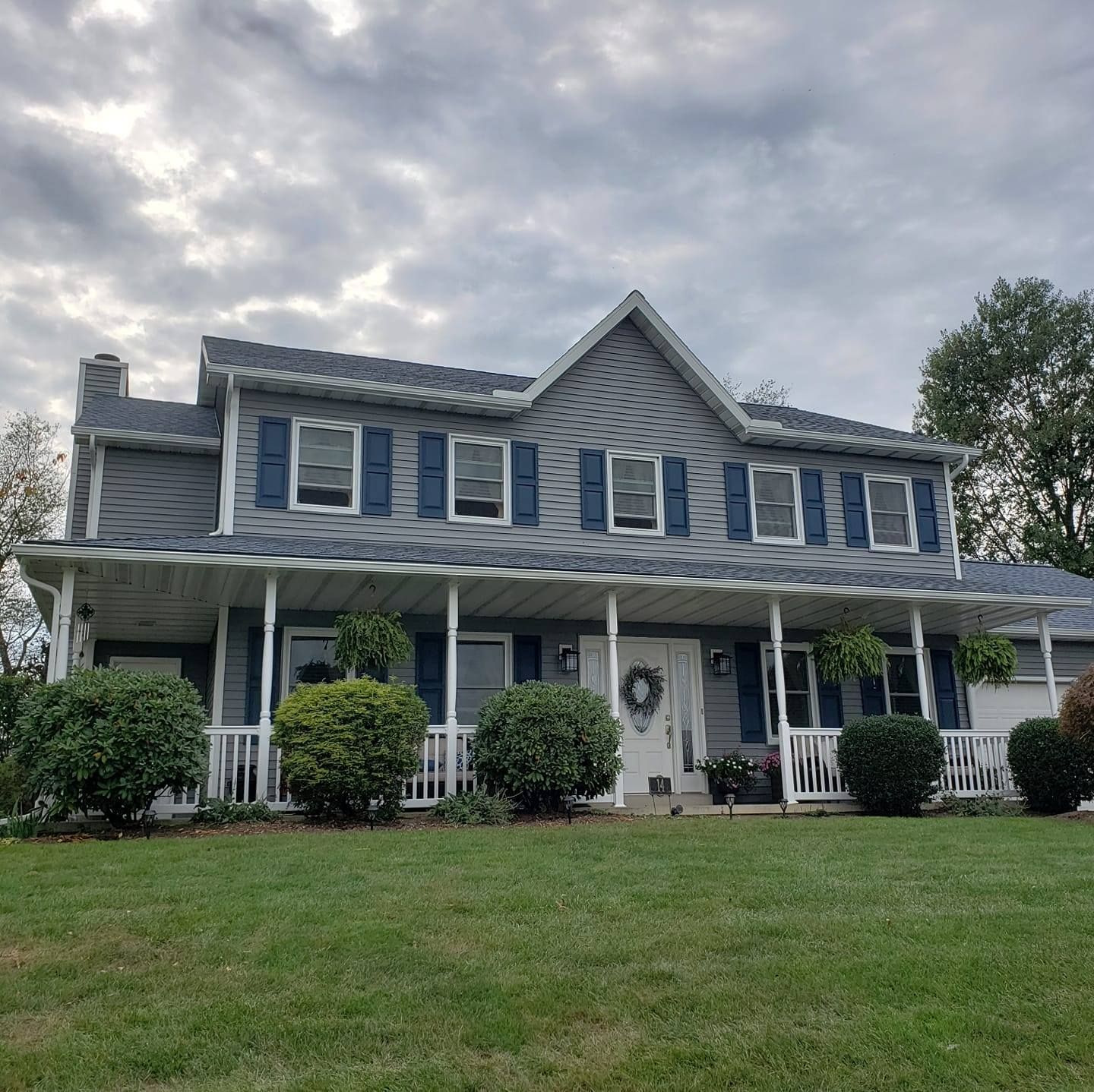 A two-story grey house with a white front porch, dark blue shutters, and green bushes under a cloudy sky.