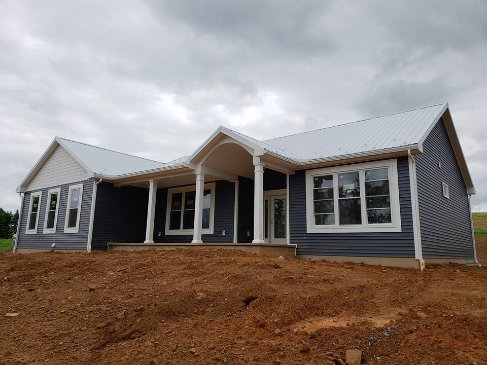 A newly built, one-story house with dark blue siding, a light metal roof, and a covered porch on a dirt construction site.