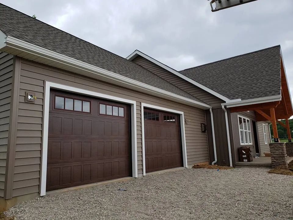 Two-car garage with brown doors and gray siding, viewed from a gravel driveway.