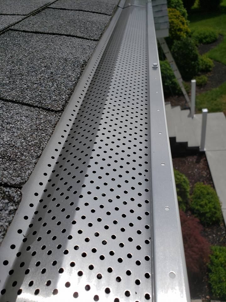 A metal gutter cover with a perforated pattern installed over a roof gutter, with a landscaped yard in the background.