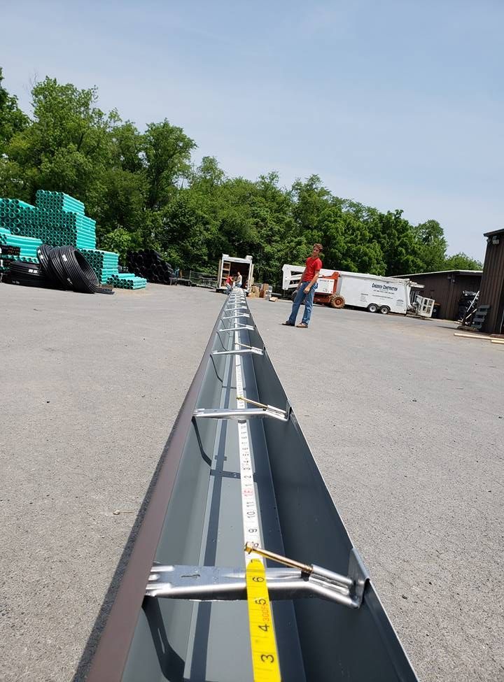 A person stands near a long, dark gutter section lying on pavement, with a yellow tape measure running along its length.