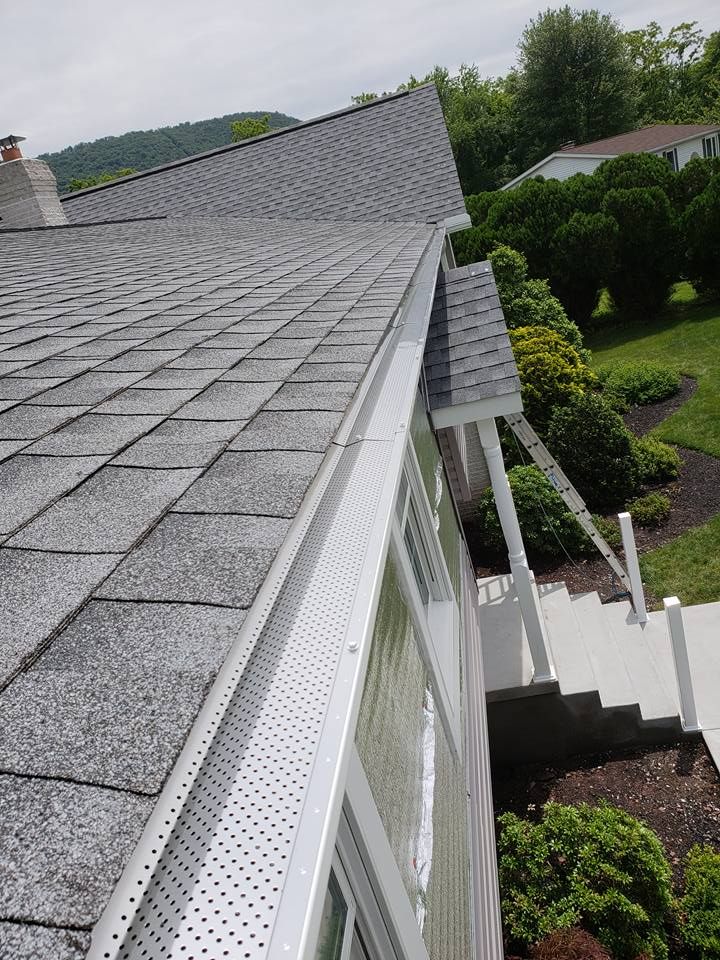 A high-angle view of a gray shingled roof with a white gutter guard installed along the edge, overlooking a front yard.