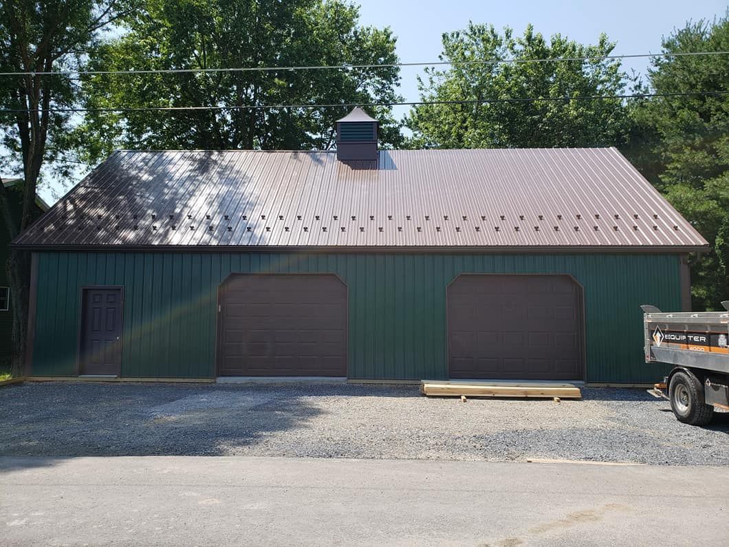 A green metal garage with a brown metal roof, two large garage doors, and a single side door on a gravel lot.