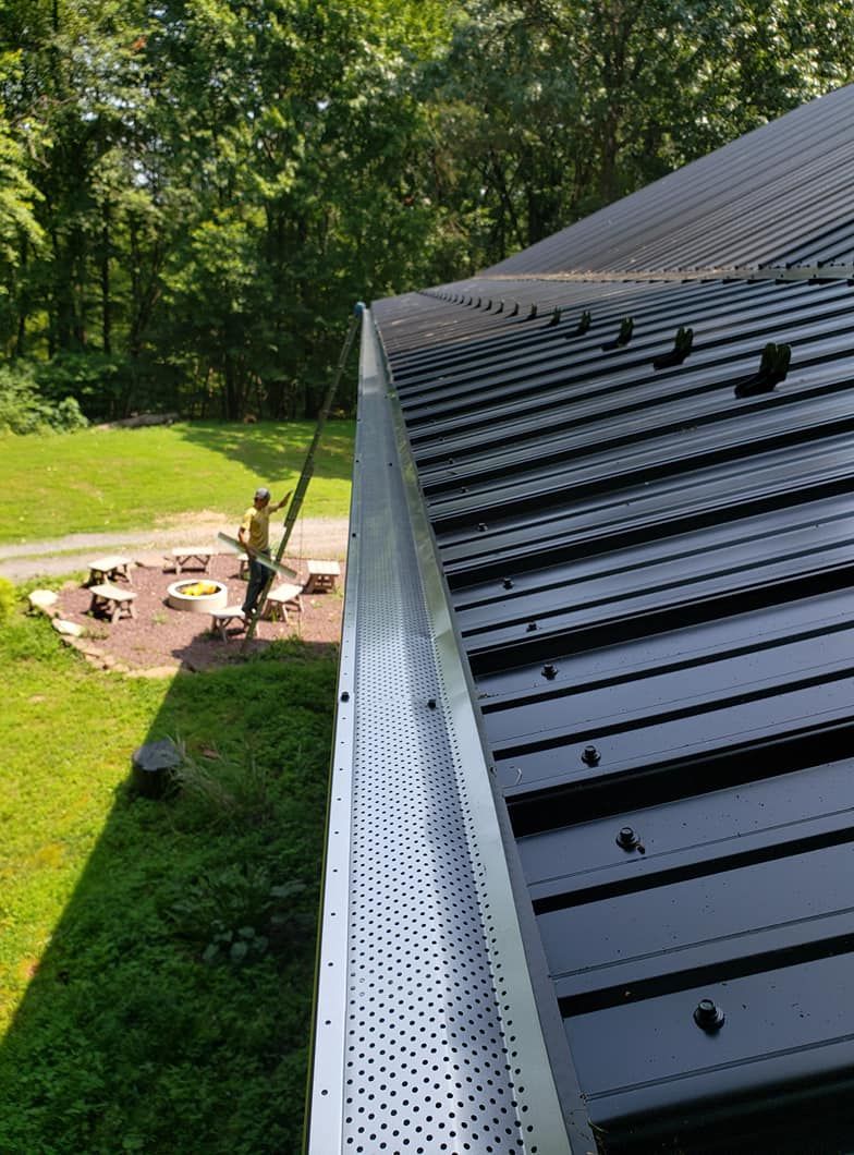 A high-angle view looking down a metal roof with gutter guards, while a person on a ladder works on the roof nearby.