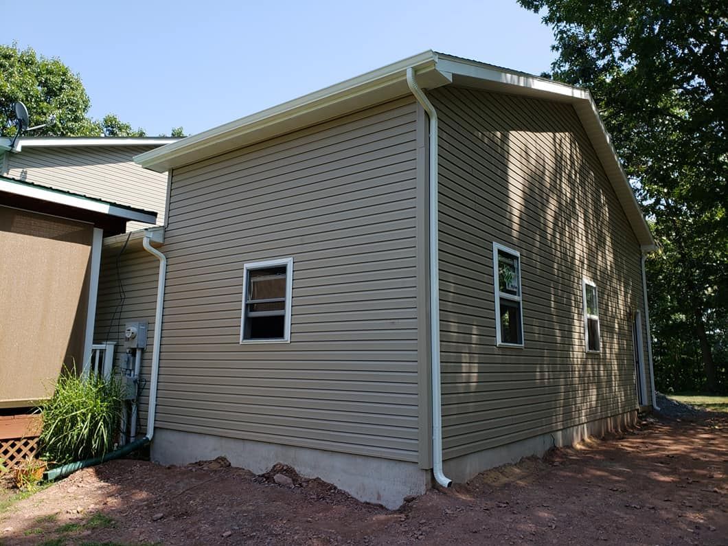 A tan vinyl-sided house with white trim and downspouts, featuring two visible windows on a gravel lot near trees.