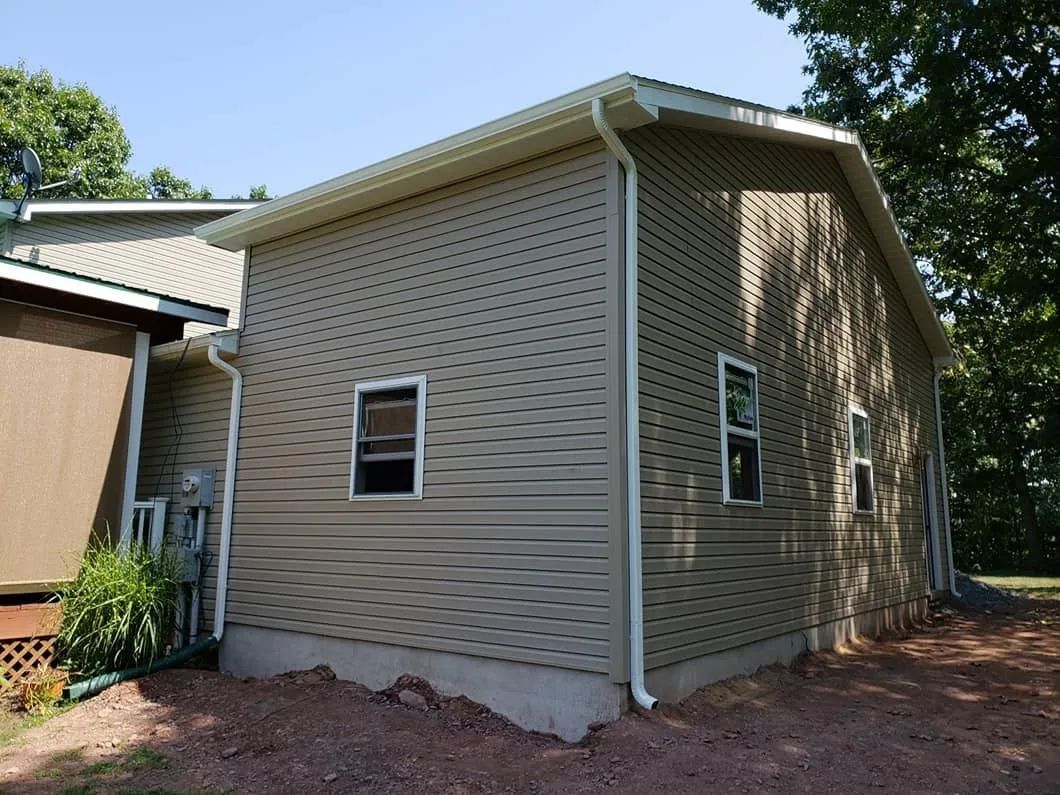 Tan house siding and roof with small windows beside a wooded yard