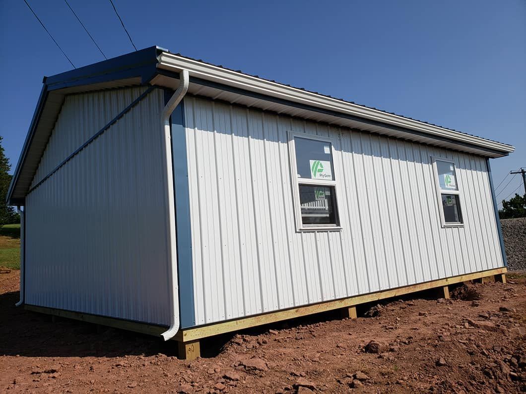 A white, metal-sided rectangular structure with a gable roof and two windows sits on a wooden foundation in a dirt lot.