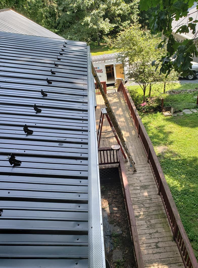 A metal roof with snow guards overlooks a wooden ramp and deck area next to a building in a grassy, wooded yard.