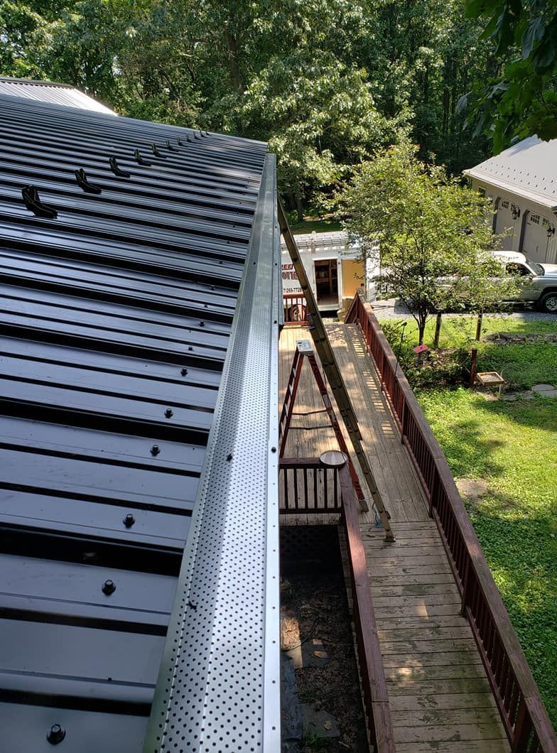 An elevated view of a metal roof edge with a gutter guard, looking down toward a wooden deck and a ladder below.
