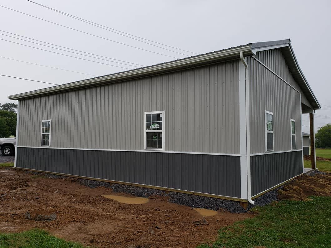 A grey and charcoal metal-sided building with two windows on a grassy, dirt-filled lot under an overcast sky.
