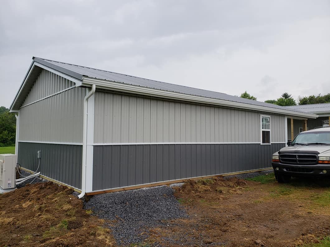 A side view of a metal building with grey walls, a white trim, and a parked truck in a grassy, partially graveled yard.