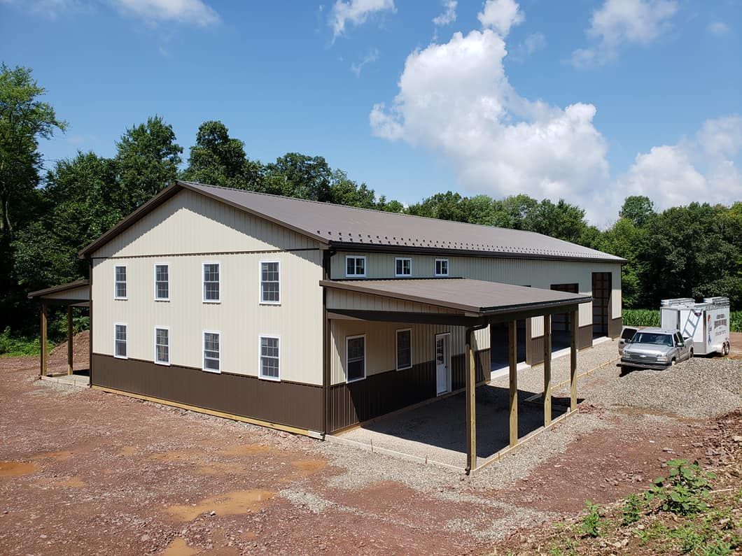 A tan and brown two-story metal building with a side porch, gravel yard, and trees under a blue sky.