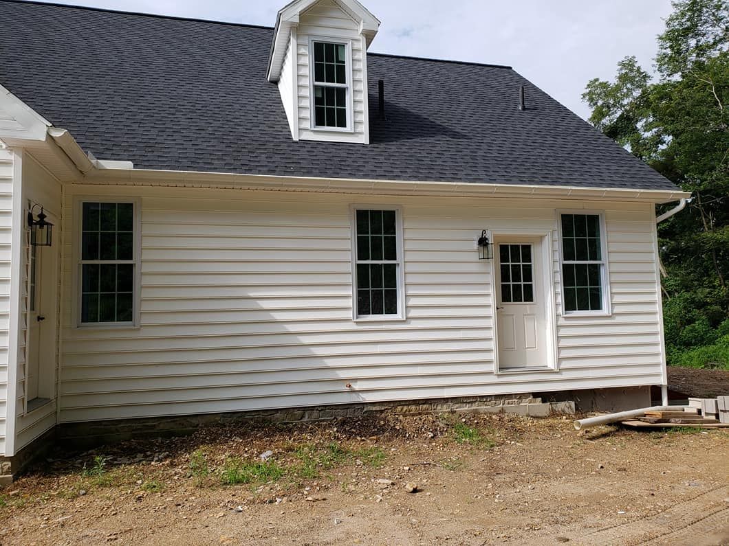 A side view of a white, clapboard-sided house with a dark shingled roof, a dormer window, and an unfinished dirt yard.