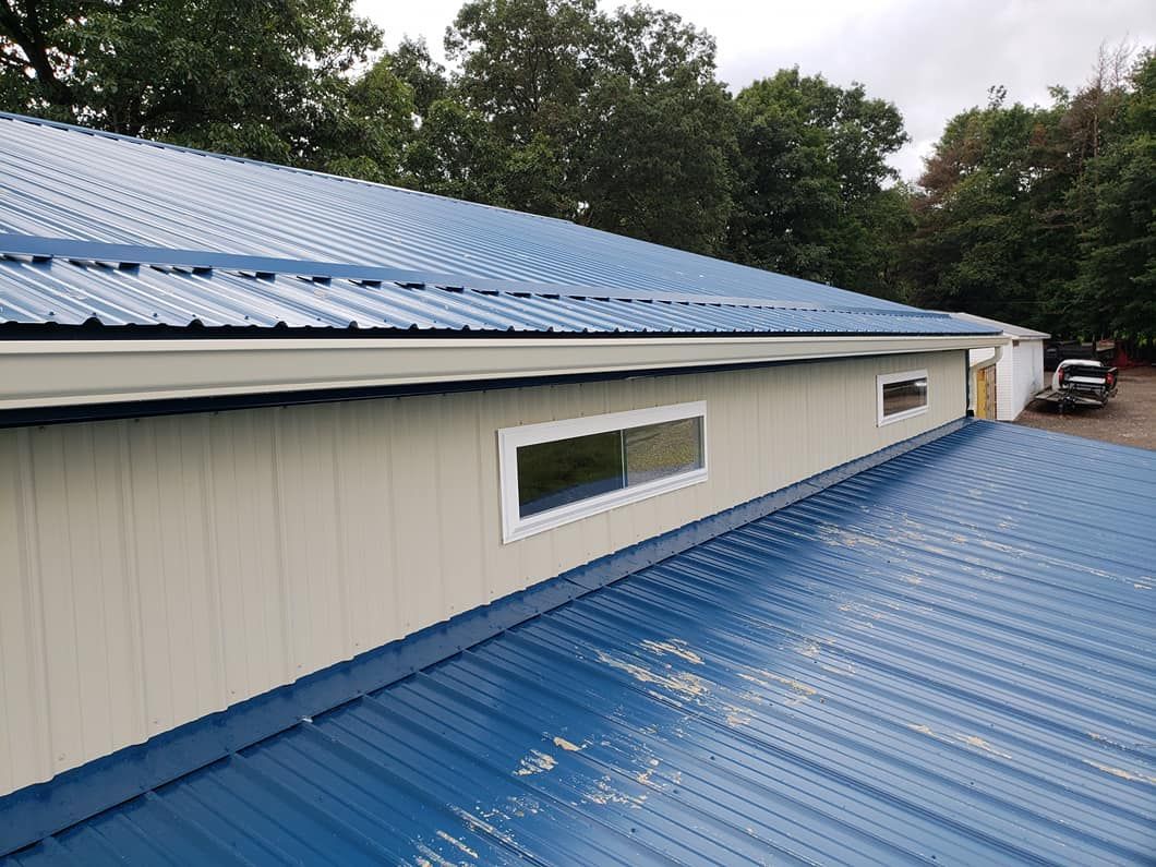 A low-angle view of a building with a blue metal roof and beige vertical siding featuring two rectangular windows.