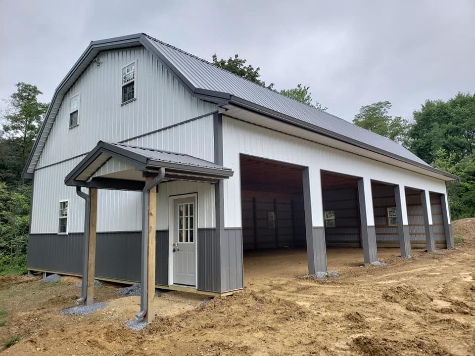A two-story pole barn with white and dark gray metal siding, a metal roof, a front porch entry, and open bay garage stalls.