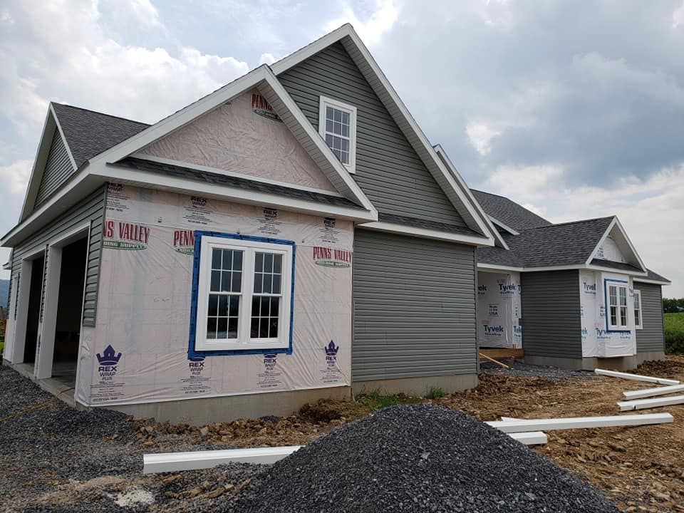 A new house under construction with grey siding, a dark roof, and white-trimmed windows, sitting on a gravel lot.