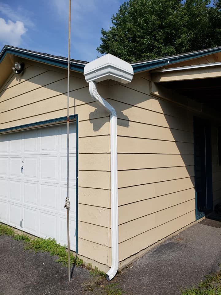 A white downspout attached to the corner of a tan house with a vertical antenna pole standing near the garage door.
