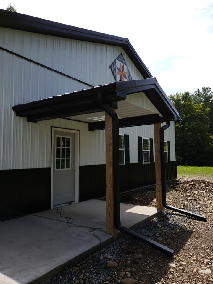 A building entrance with white siding, a black lower trim, a porch roof with brown wooden posts, and a decorative quilt.