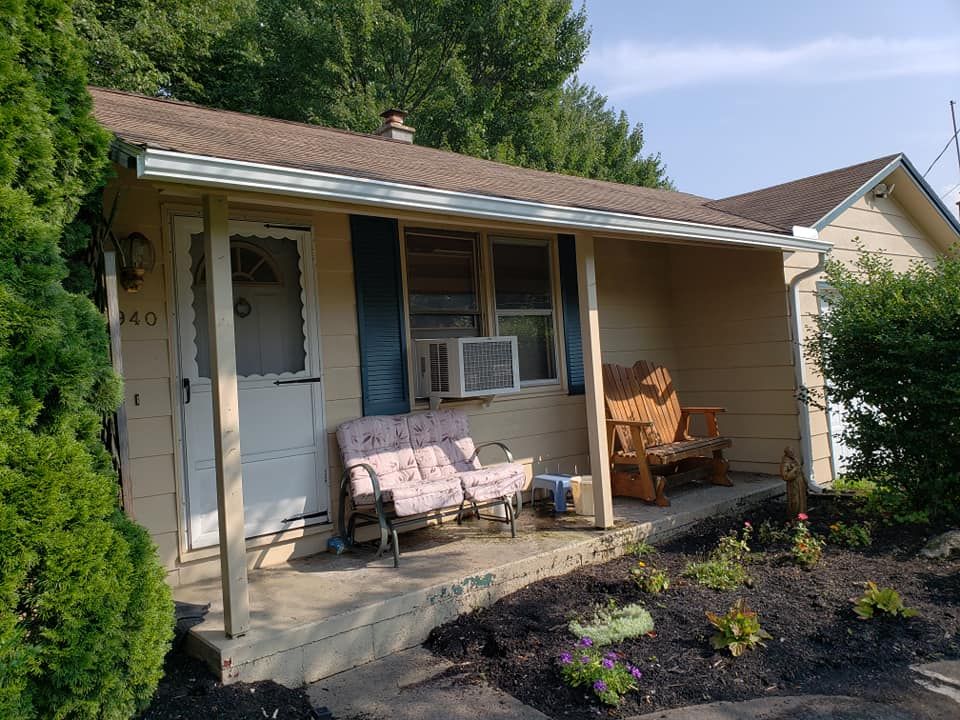 A tan-sided house with a front porch, featuring a small loveseat and a wooden bench, plus an air conditioning unit.