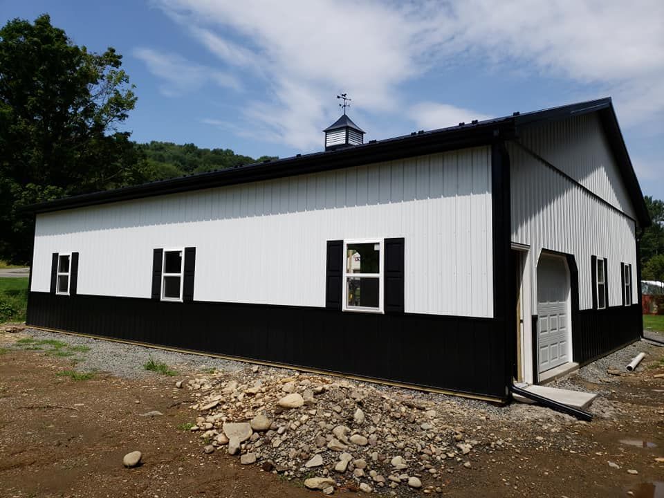 A white barn with a black lower trim, black roof, shutters, and a cupola on a sunny day with trees in the background.