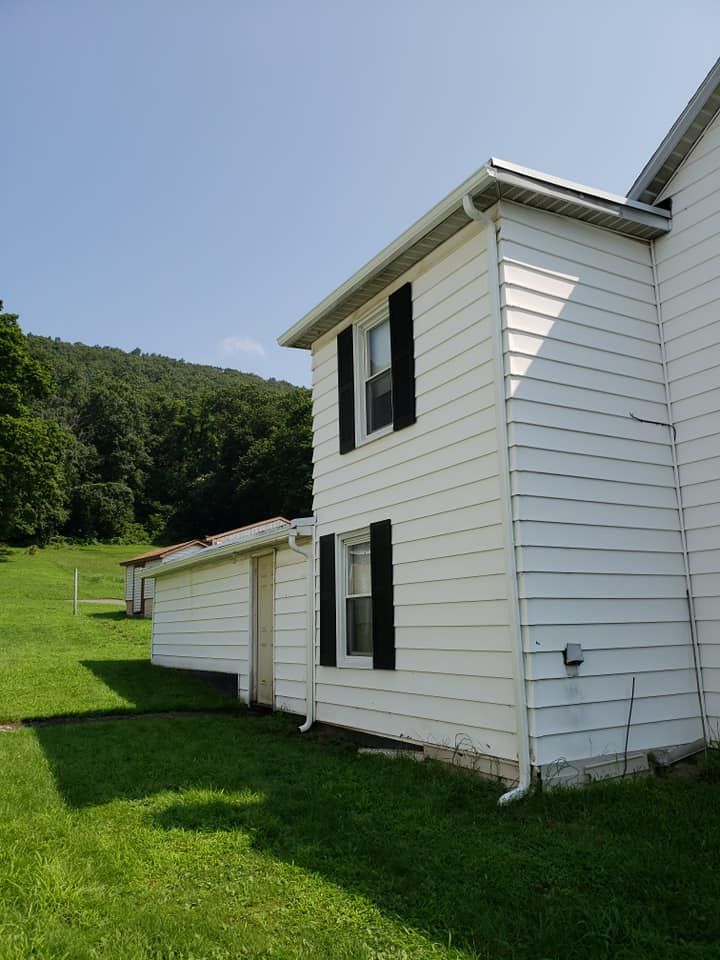 A two-story white house with black shutters sits on a grassy hill next to a forest under a clear blue sky.