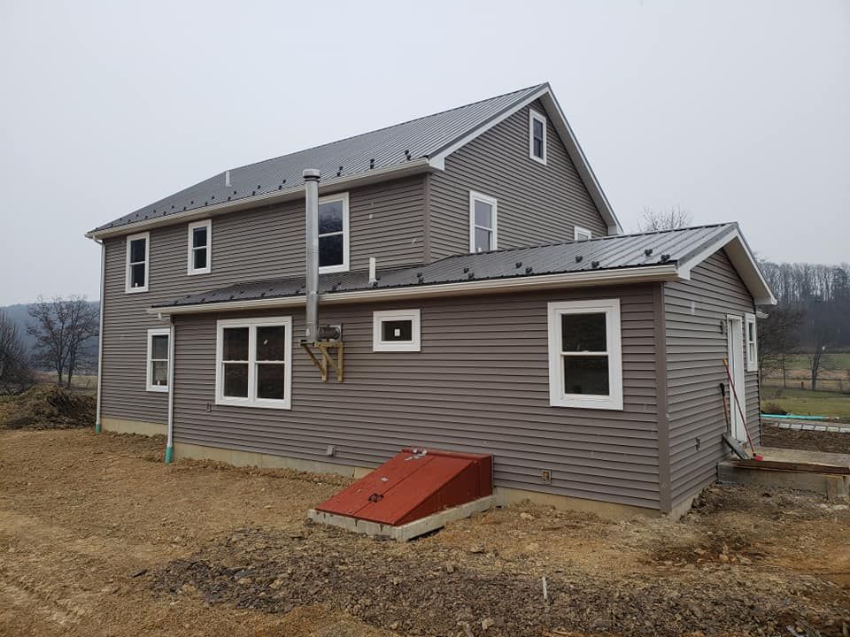 A two-story grey vinyl-sided house with a metal roof and a red bulkhead cellar door on a dirt lot under an overcast sky.