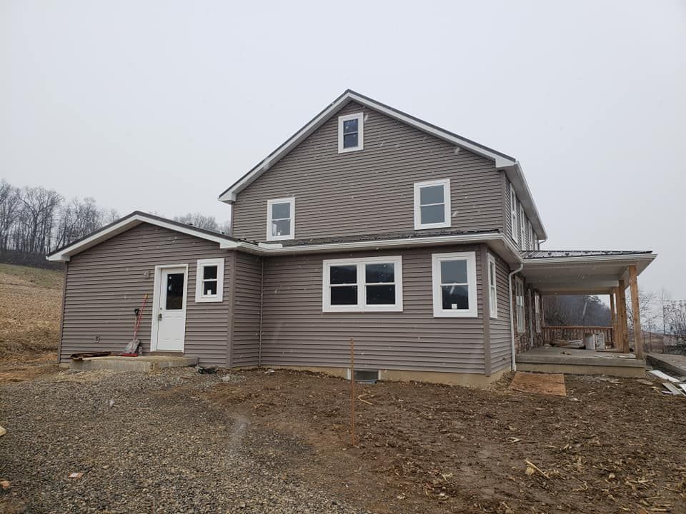 A two-story grey-sided house with a porch under construction, set in an unfinished yard on an overcast day.