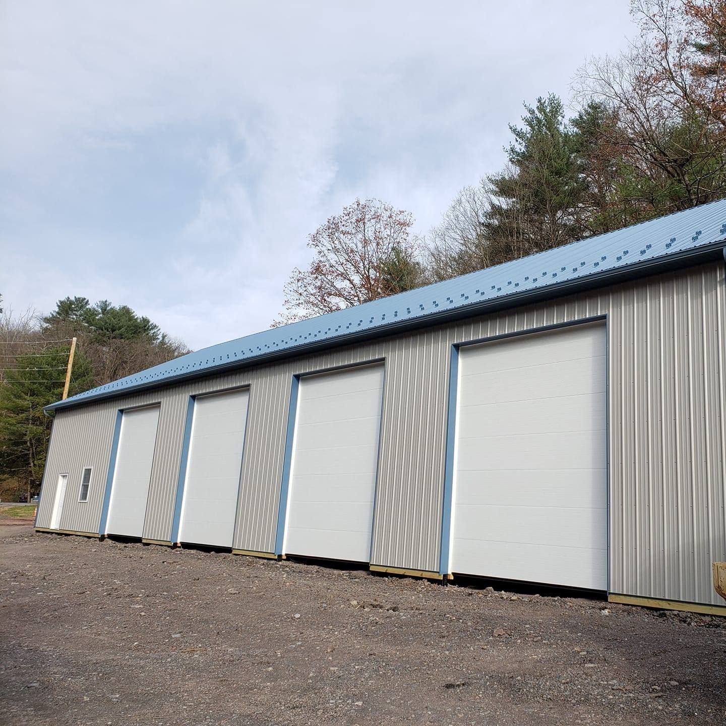 A metal storage building with four white roll-up doors, a blue roof, and a gravel parking area against a tree line.