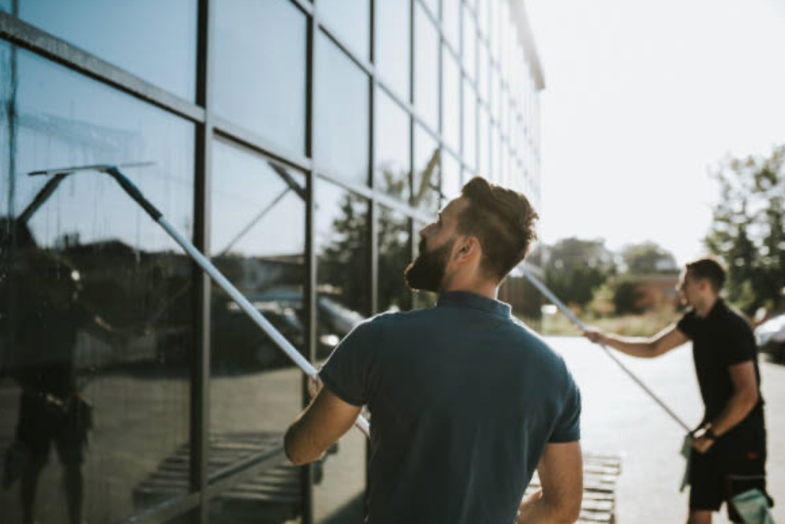 Two men washing windows of a modern building with long-handled squeegees on a sunny day.