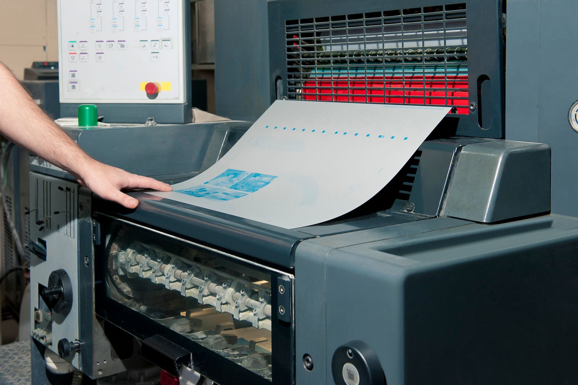 Person feeding a sheet into a printing press with red paper stacked in the background