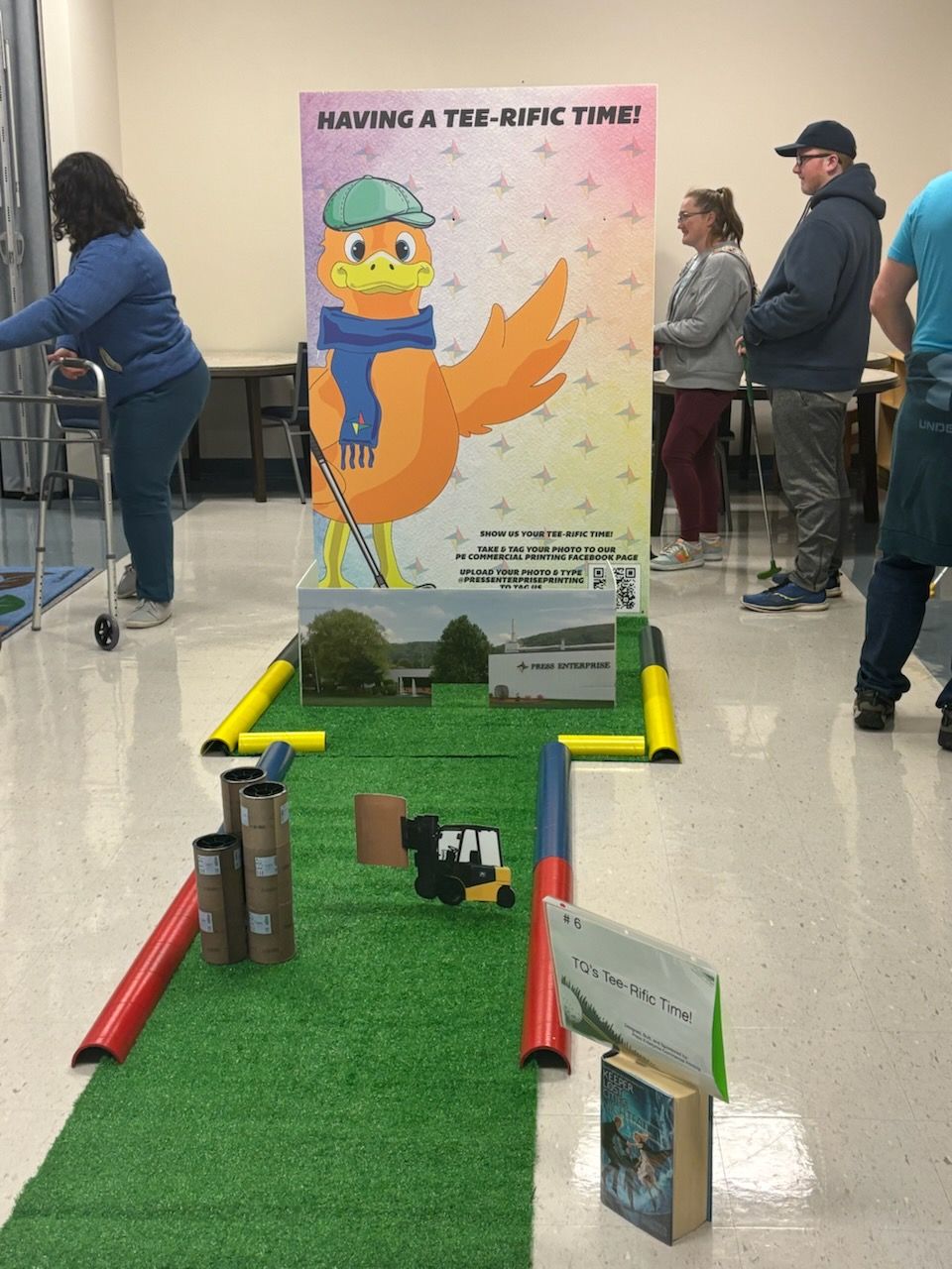 Mini golf setup with a cartoon bird sign, people watching, and a woman using a walker.
