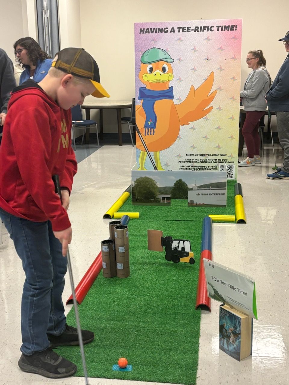 Boy playing miniature golf; green turf, cardboard obstacles, orange ball, poster of bird.