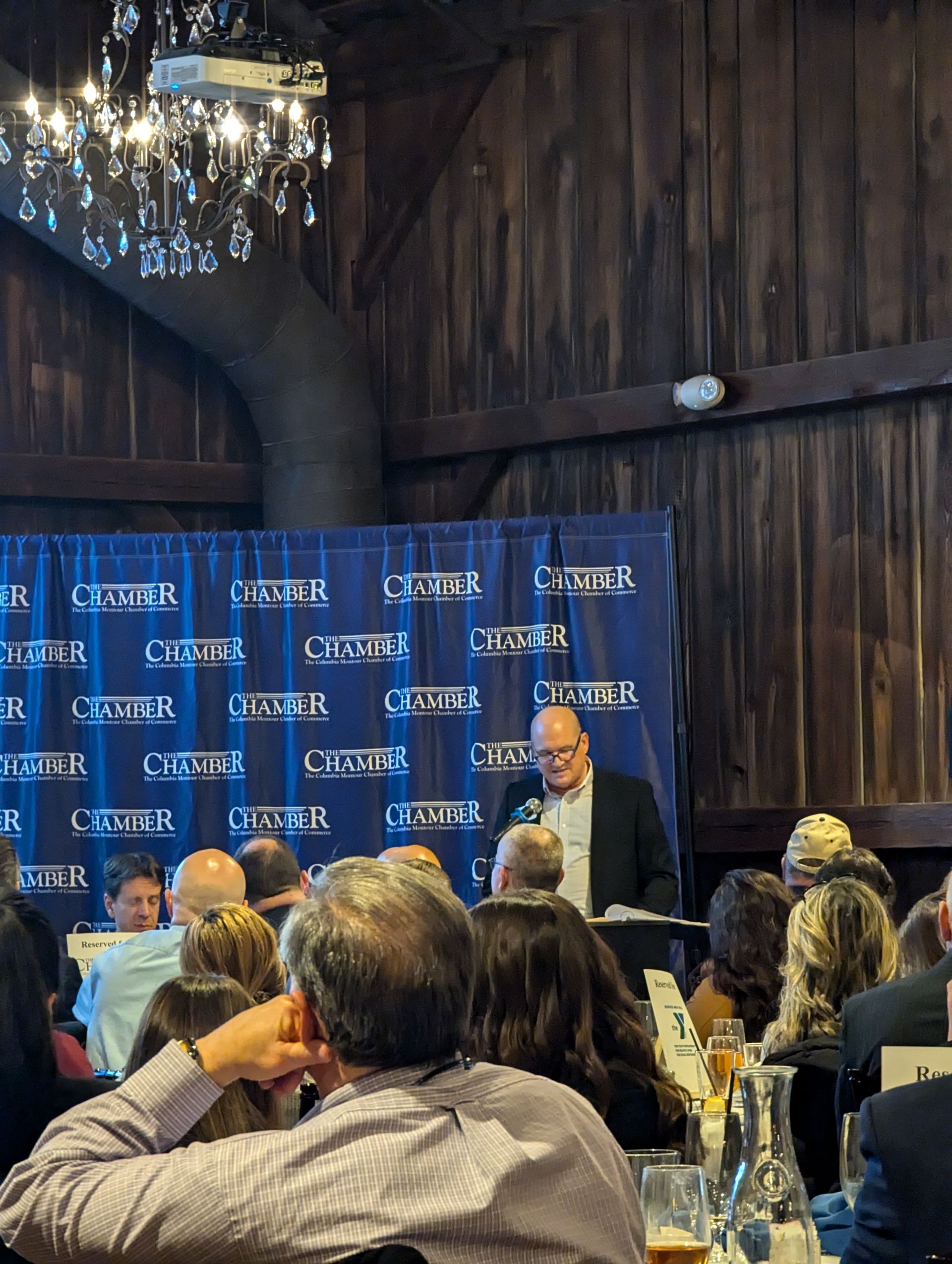 Man speaking at podium in a venue, event banner, audience seated at tables.