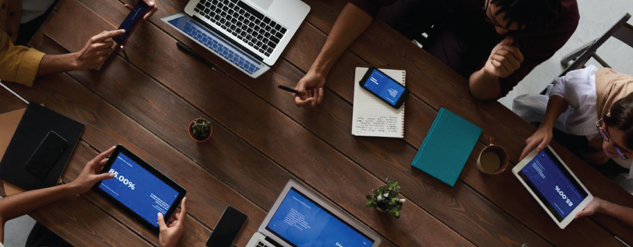 A group of people are sitting around a table with laptops , tablets , and phones. A group of people are sitting around a table with laptops , tablets , and phones.