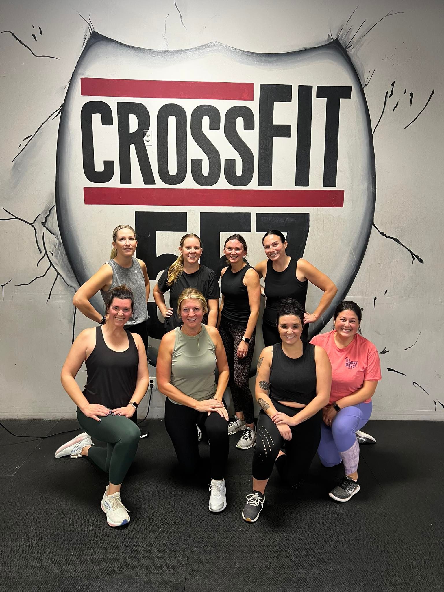Eight women posing together in workout clothes against a gray wall, smiling in a gym setting