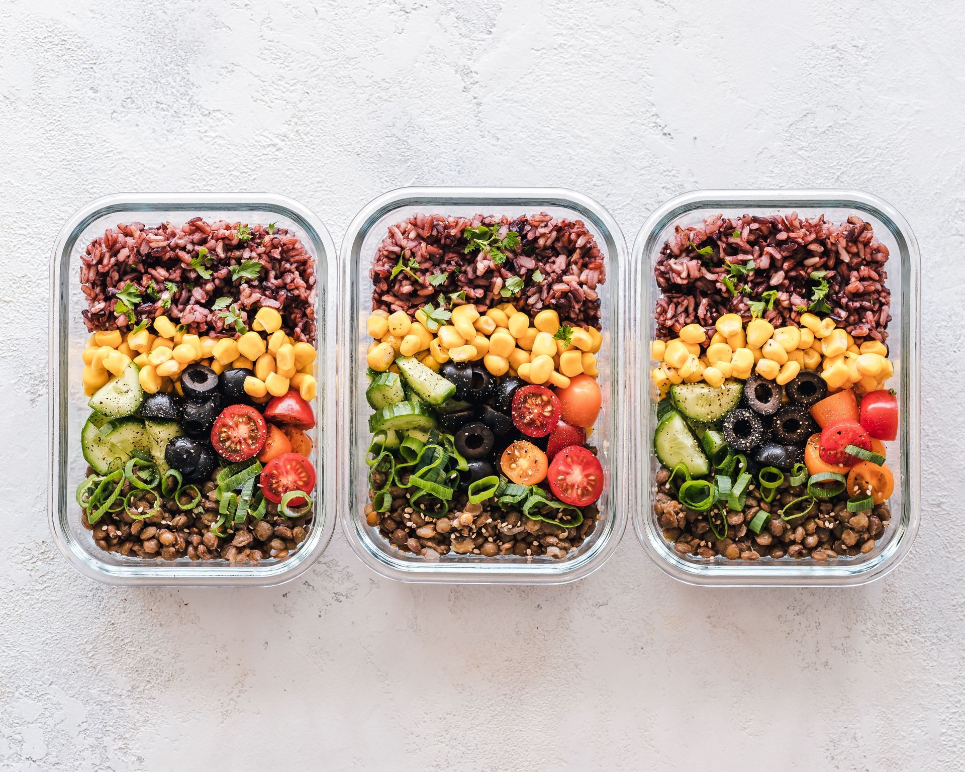 Three glass meal-prep containers filled with colorful salads, grains, berries, and vegetables on a white surface