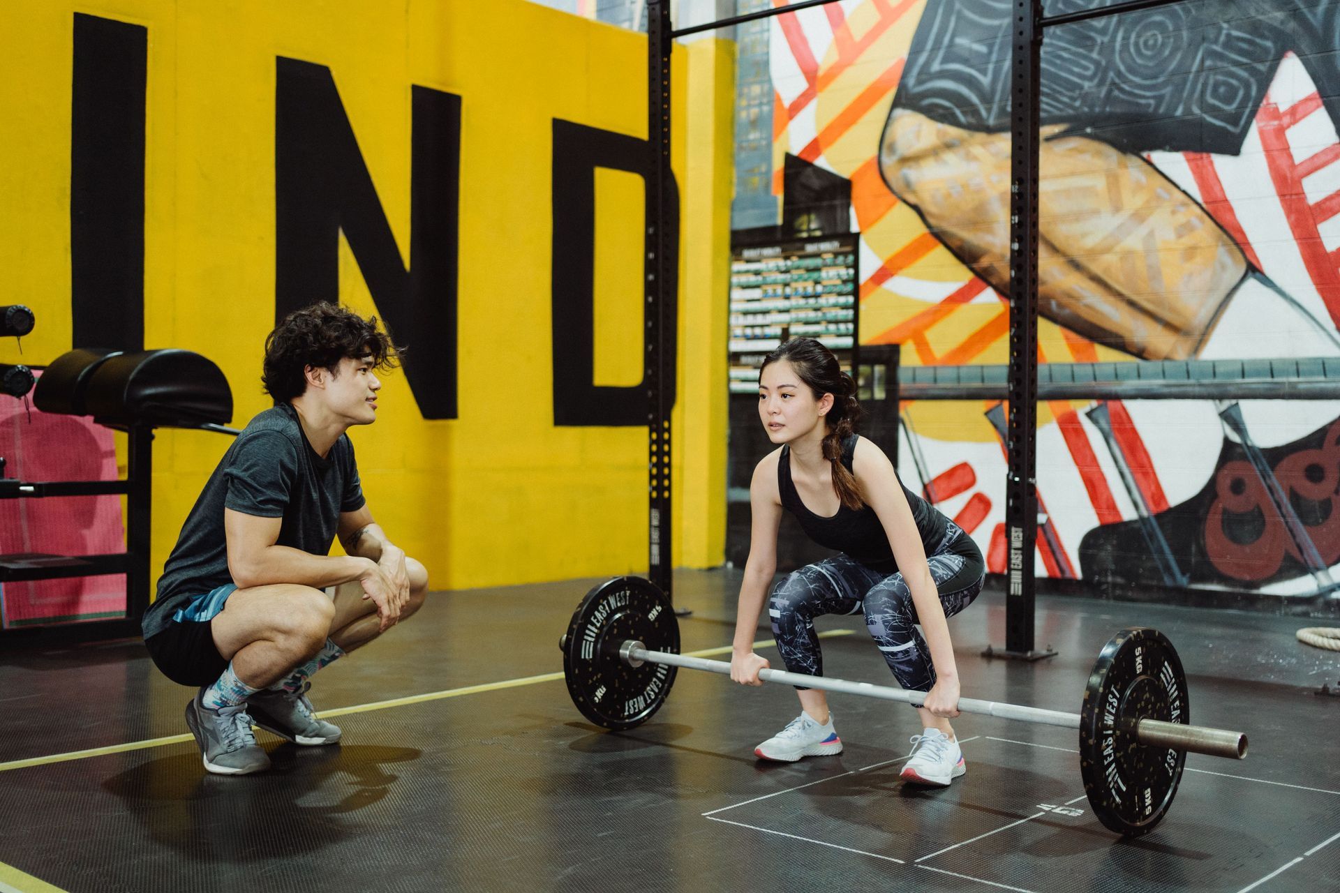 Two people crouch facing each other with a barbell in a colorful gym mural background.