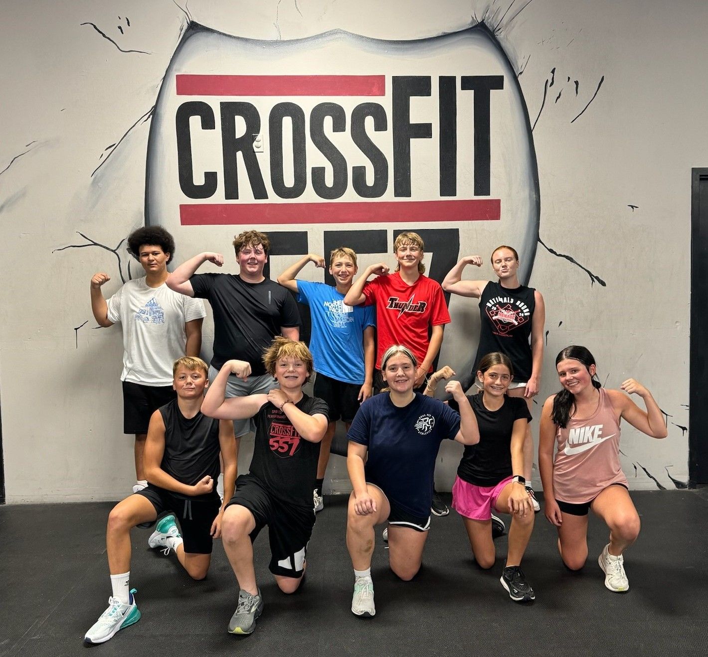 Group posing and flexing in a CrossFit gym in front of a large CROSSFIT wall sign.