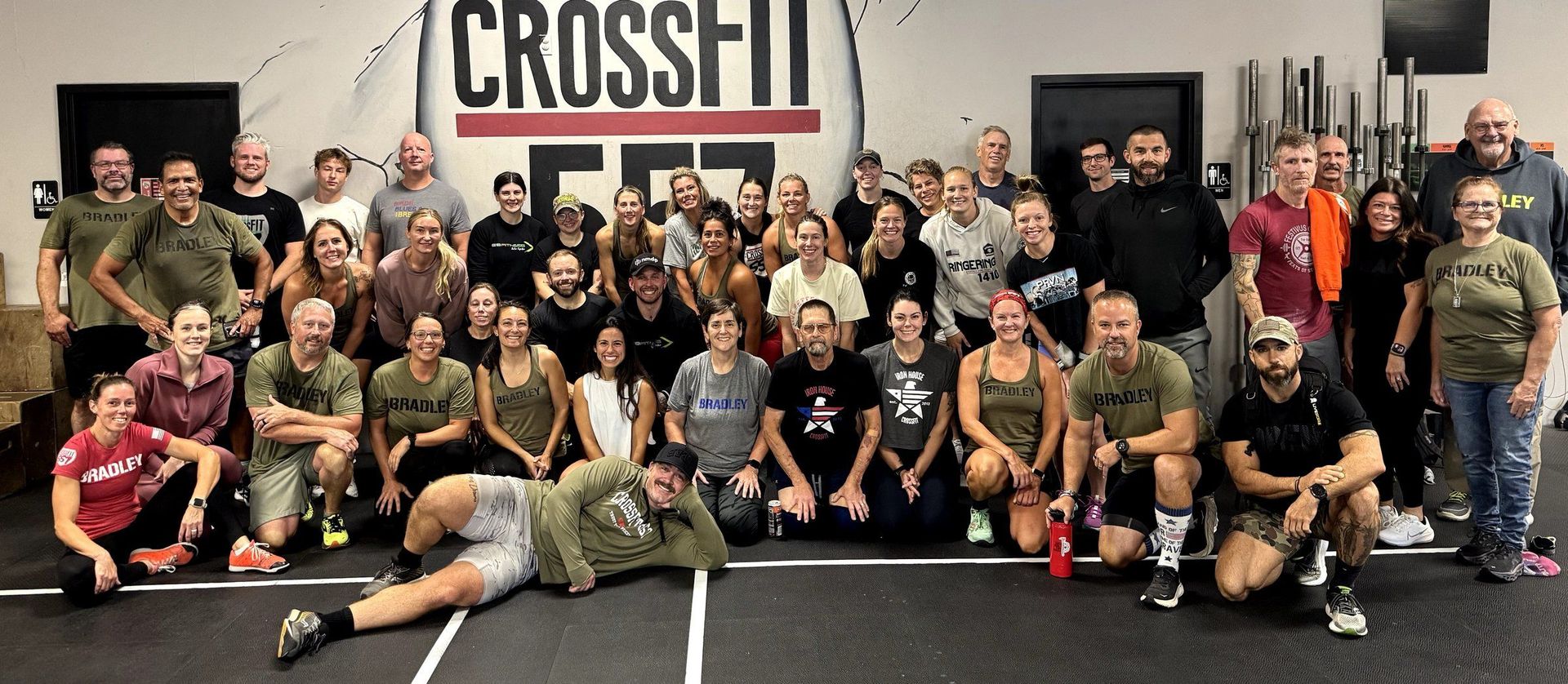 Large group posing in a CrossFit gym, standing and kneeling in front of a wall sign.