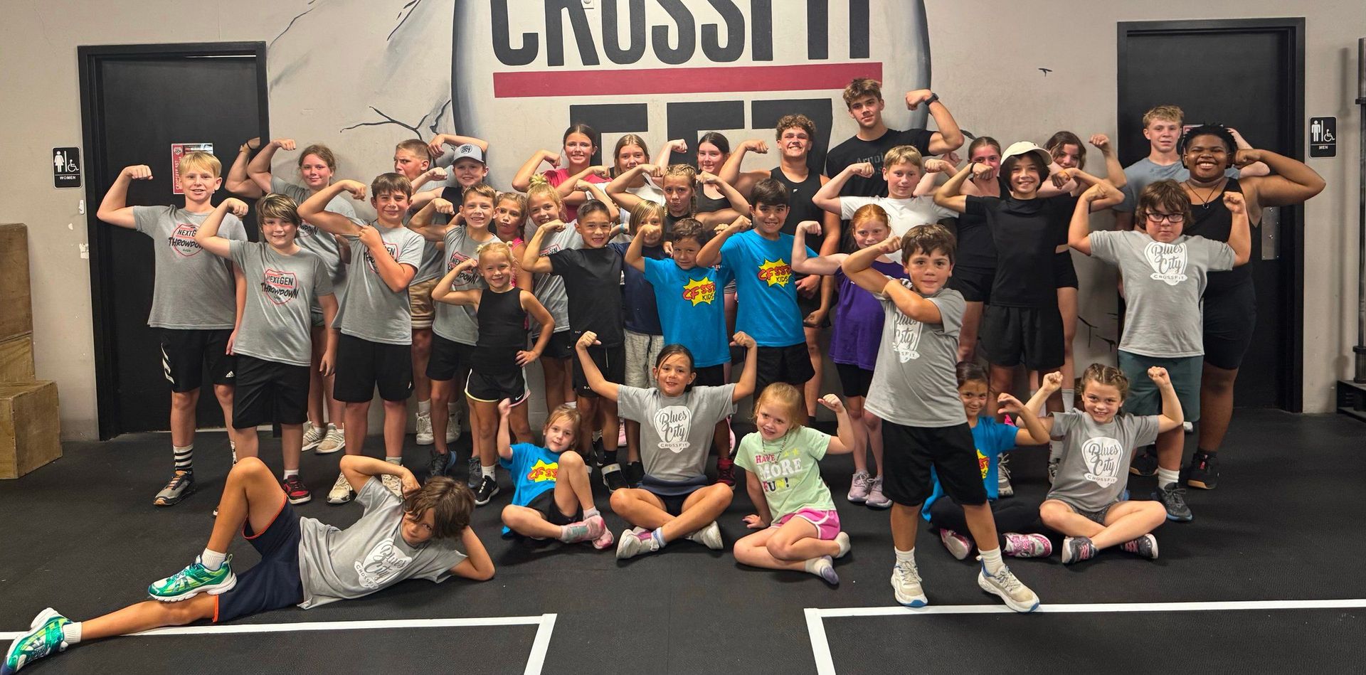 Group of people posing inside a CrossFit gym, many flexing and smiling in front of a wall sign