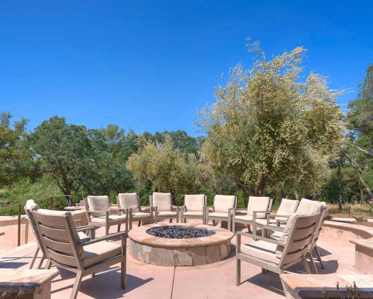 Outdoor patio with a large circular fire pit, cushioned chairs, and trees under a clear blue sky