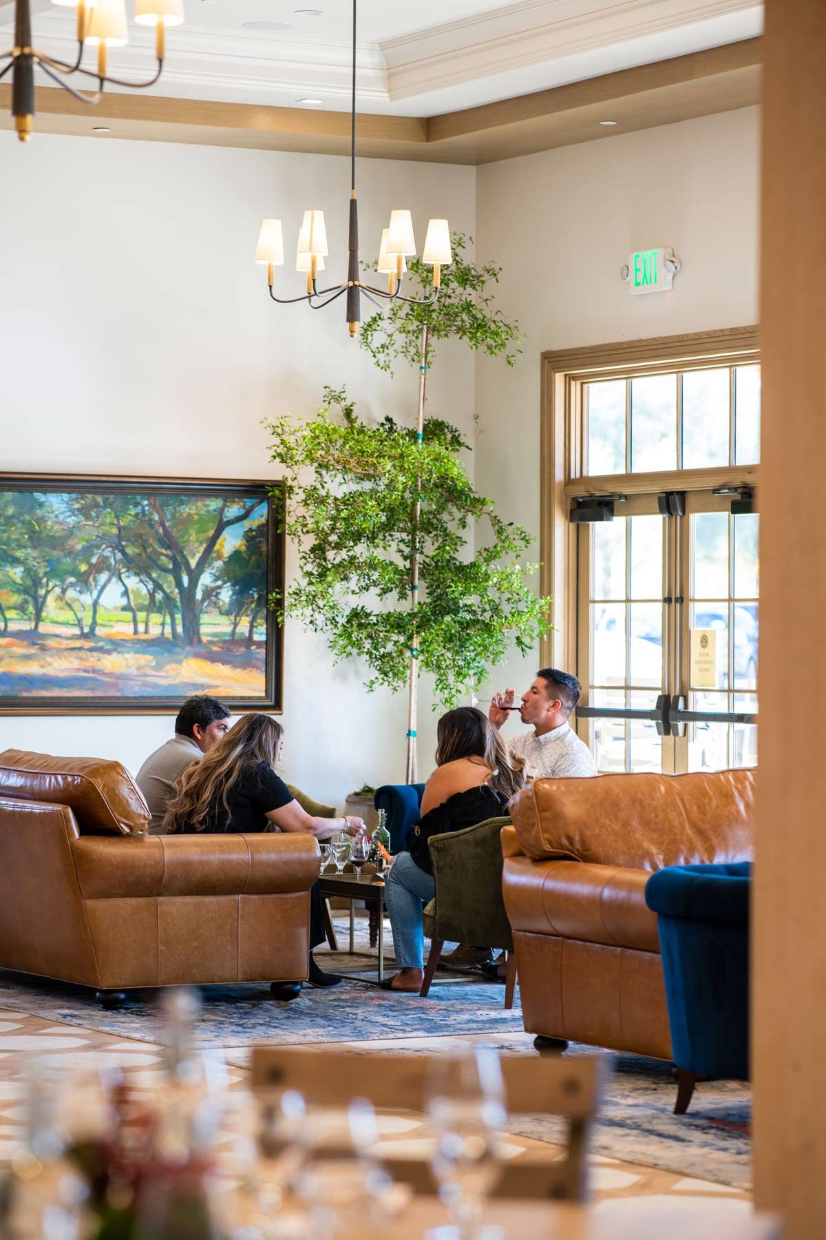 People seated in a bright lobby lounge with leather chairs, tall windows, and a large wall-mounted TV.