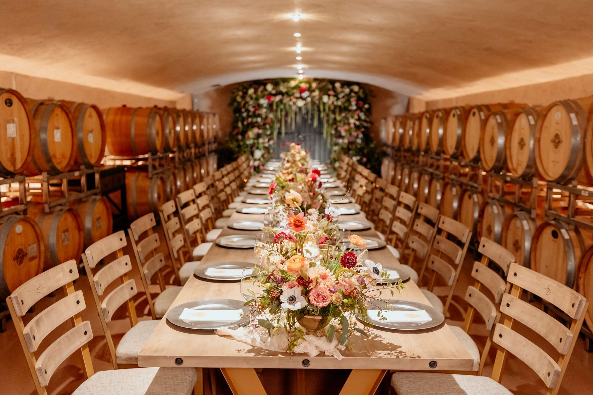Long banquet table in a wine cellar with wooden barrels, floral centerpieces, and place settings