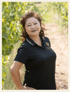 Woman in a navy polo shirt standing outdoors on a sunlit path, smiling with hands on hips.