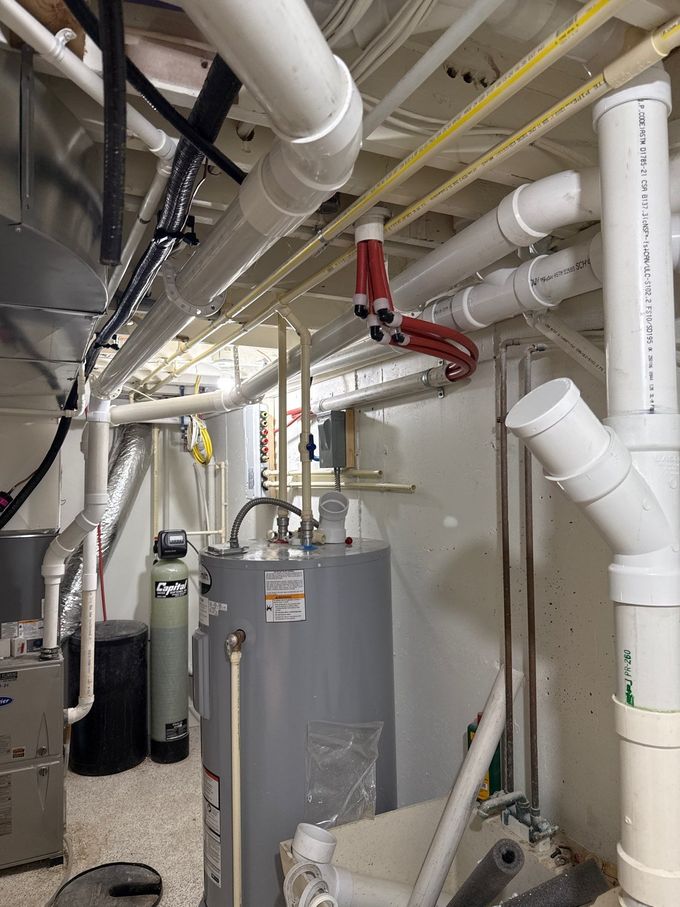 Technician in plaid shirt adjusts ceiling pipes beside a white tank in a utility room.