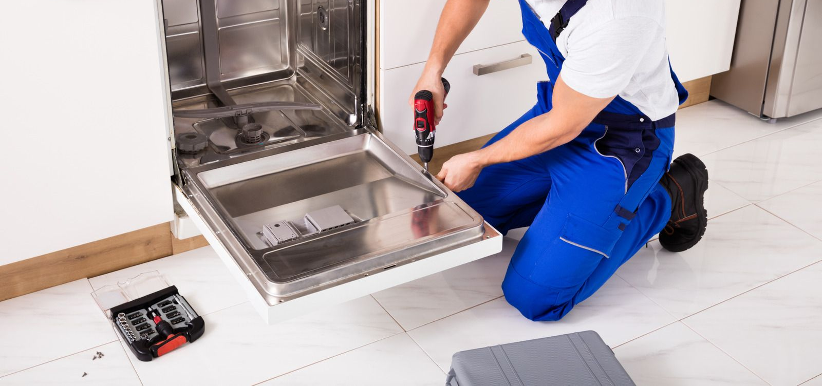 Technician kneeling beside an open stainless steel appliance, using a power drill for repair.