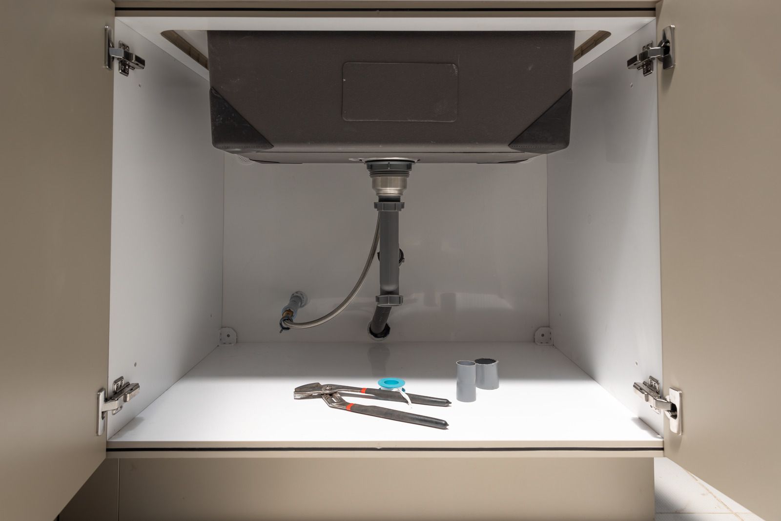 Open cabinet sink plumbing with tools and a flashlight on the white shelf below.