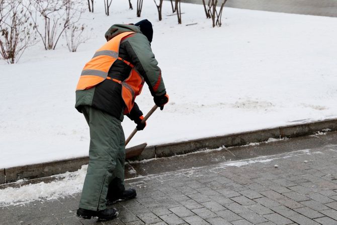 Worker in orange safety vest shoveling snow from a sidewalk near a curb