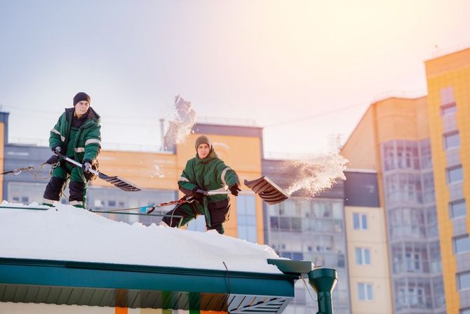 Two workers in green jackets removing snow from a rooftop at sunset